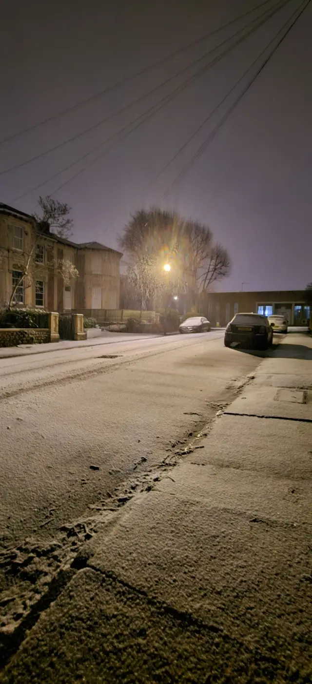 A light layer of snow covering a street in Clifton. It is dark, with streetlamps for light and a couple of cars at the end of the road