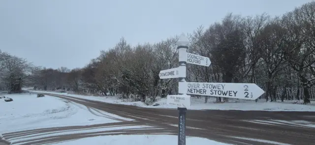 An old-fashioned road sign at a cross-roads. The road is covered in snow, with dark tyre marks running though it. There are lots of trees in the background, all bare with snow covering them.