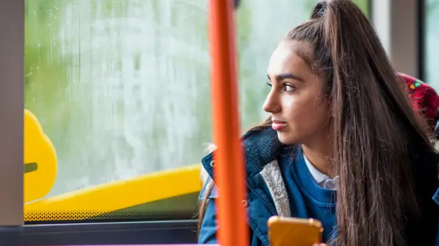 A teenage girl with long brown hair in a high ponytail sitting on a bus and looking out a steamed-up window. She is holding her phone in her hand.