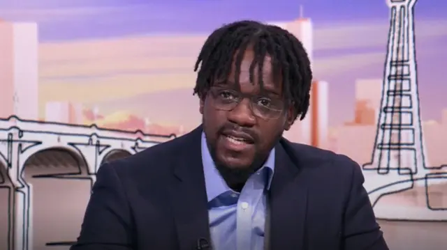 Samuel Kasumu sitting down at desk during panel appearance. He's wearing a dark suit with a blue shirt, no tie. In the background is a sketched cardboard illustration of UK landmarks in pastel colours