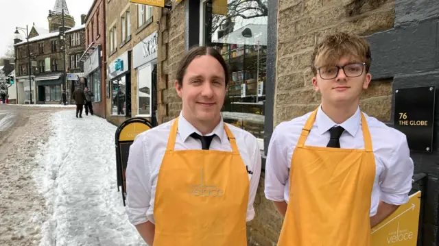 Anthony Black and Oliver Wilde stand with their arms behind their backs wearing yellow aprons outside their cafe. Snow can be seen in the street.