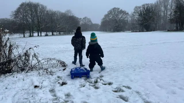 Two children bundled up in snowsuits or thick coat and wellies stand with a blue sledge on a gentle snow-covered slope on a golf course.