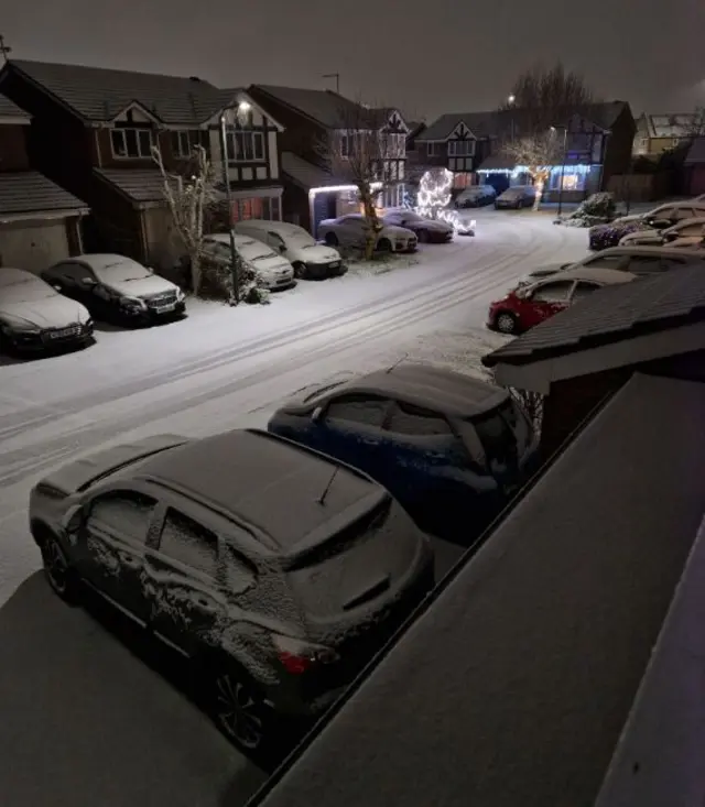 A street of houses, with cars parked outside, covered in a light dusting of snow. There are car tracks in the middle of the road.