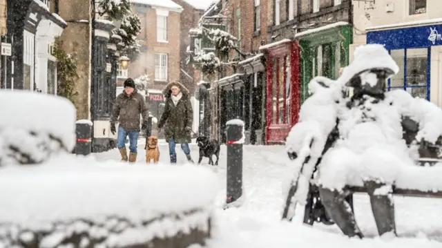 A man and a woman walk two dogs (one brown walking between then, one black walking next to the woman's left) through the snowy streets of Knaresborough, Yorkshire. Closed shops and homes to the side of the road