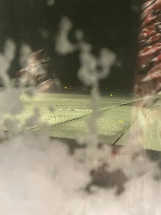 A plane window frosted over with a snow-covered run way seen in the distance at night.