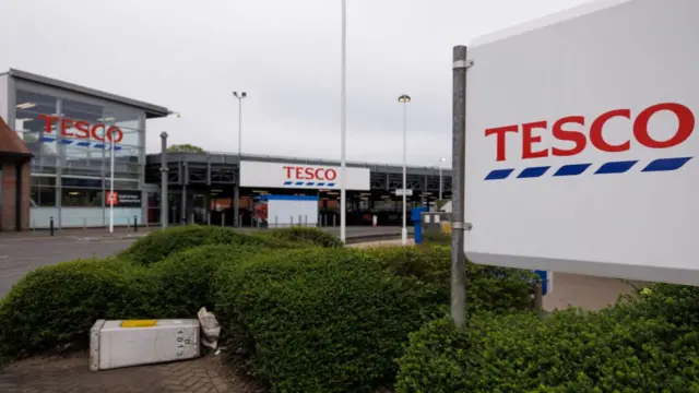 A general view of a Tesco store in London. In the foreground, is a board with the Tesco logo (white background, Tesco in red lettering, underline by blue dotted line) with underneath green bushes. In the background is a superstore marked Tesco both at the main entrance and to the side