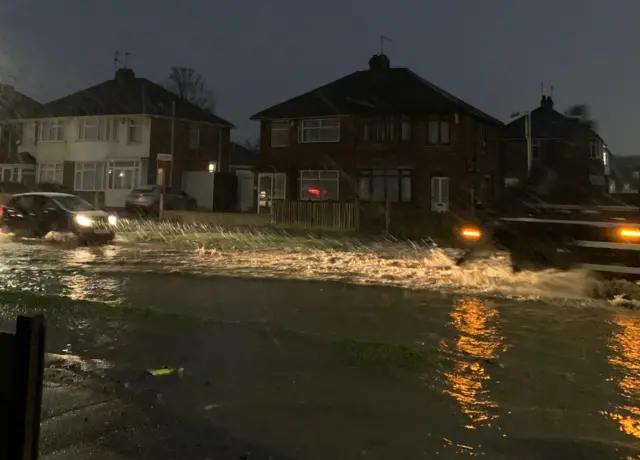 A flooded road at night with cars driving through leaving trails in theor wake