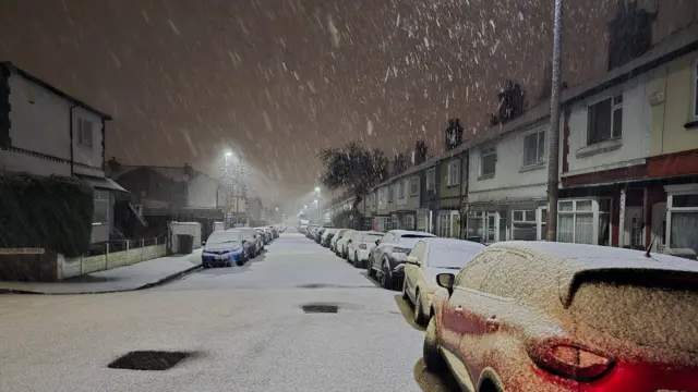 A snow covered road and snow falling from the sky. There are rows of houses on either side of the street, as well as a row of cars on either side. The cars are covered in snow.