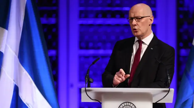 First Minister John Swinney giving a speech at Playfair Library in Edinburgh, at a lecturn with microphones and by a Saltire