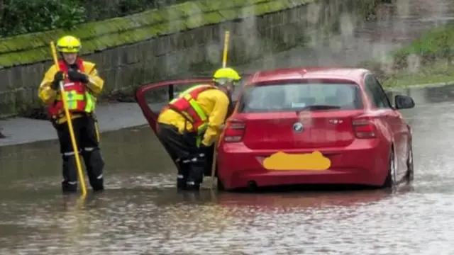 Two firefighters who are standing in floodwater by a red car which has stopped in the water