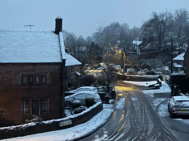 A snow-covered village with a road going through it. The road has tyre marks on it and melted snow. Trees can be seen in the distance with houses and parked cars either side of the road.