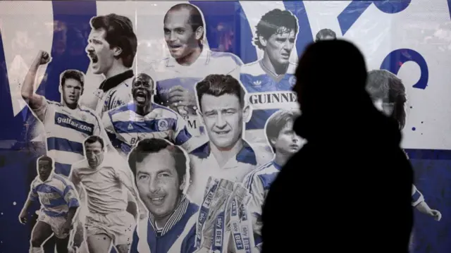 A silhouetted fan walks past a mural dedicated to QPR legends outside Loftus Road