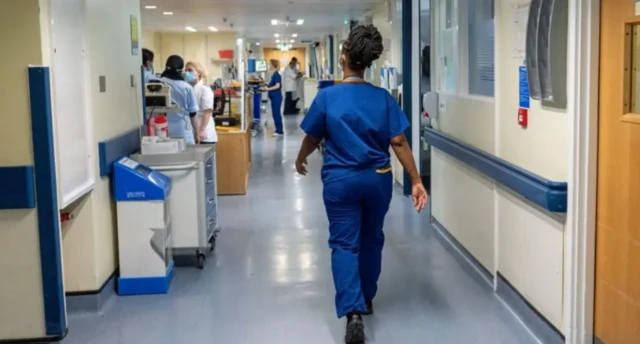 An NHS staff member in blue overalls walking through a hospital corridor with her back facing the camera