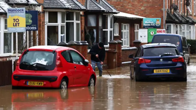 A man poking a stick in flood water, which goes above his ankles. He stands on a flooded residential street, with three cars sitting in the flood