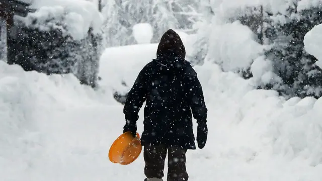 A woman in a dark coat with the hood up, her face obscured, walks on an icy road after heavy snowfall holding an orange plastic tub