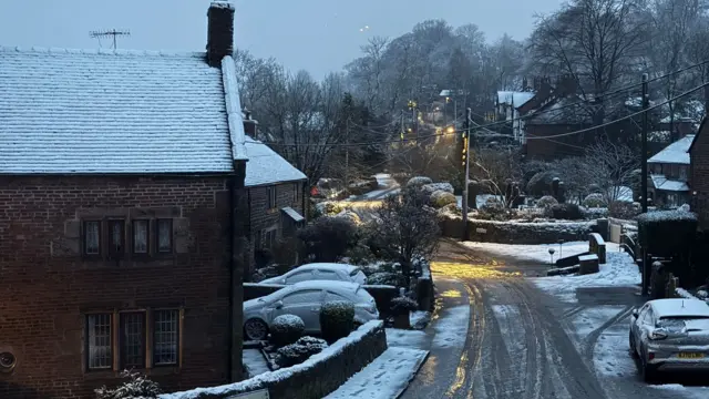 A snow-covered village with houses and cars either side of a road, which is covered in melted snow. Trees can be scene in the background as the road curves to the left.