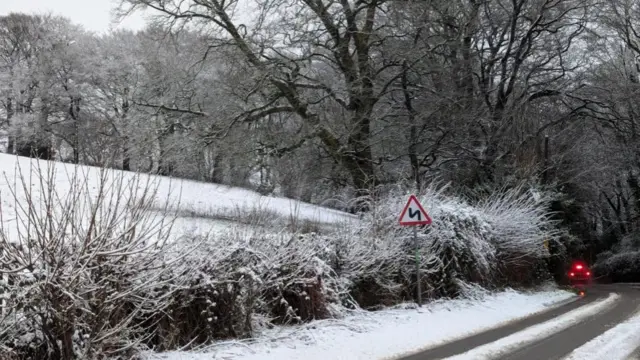 A car goes around a corner on a snow-covered rural road, with the adjacent hedge, field and surrounding trees also covered in snow. A warning sign is also visible on the left.