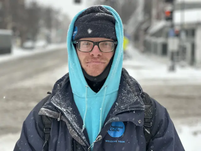 A man in a big blue coat saying Dupont Circle with a light blue hoodie, with hood up, wearing a black hat and snood with snow in the background