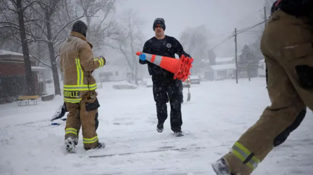 Firefighters work to clear snow on road, two in brown and yellow outfits walking by as man in black outfit holds an orange traffic cone in his hands