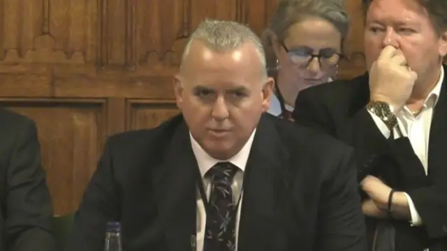 McDonalds CEO Alistair Macrow in black suit, white tie and patterned tie sits at wooden table while giving evidence to the Commons Business Committee. Behind him, to his left, is a man in a black suit, large watch on his right wrist and a black wristband on his left wrist; a woman in glasses looking to a phone (reflection visible in the glasses)