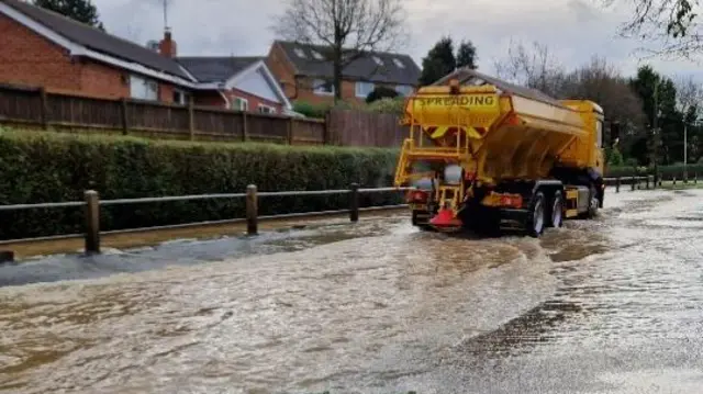 A yellow gritting lorry passing along a flooded road