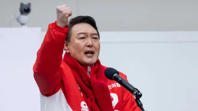 Yoon Suk Yeol in a red scarf and jacket raising his fist while speaking at a rally