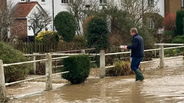 Man holding tea mugs whilst walking through flood water in his wellies