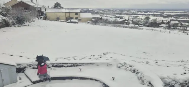 A view over the countryside covered in snow. A child can be seen petting a pony next to a wall in the foreground.