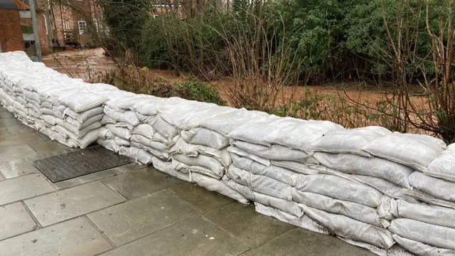 A makeshift wall of white sandbags piled up in front of a brook which is brown. There are bushes and shrubbery either side of the water, which runs into the distance near some buildings