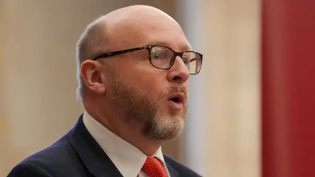 Close up shot of Labour MP Liam Byrne, cutting off just below the neck. He's wearing thin-rimmed black glasses, a dark blue suit jacket and a red polka-dotted tie. Photo is of the right side of his face as he looks straight ahead while speaking, in the background is a red wall with white accents