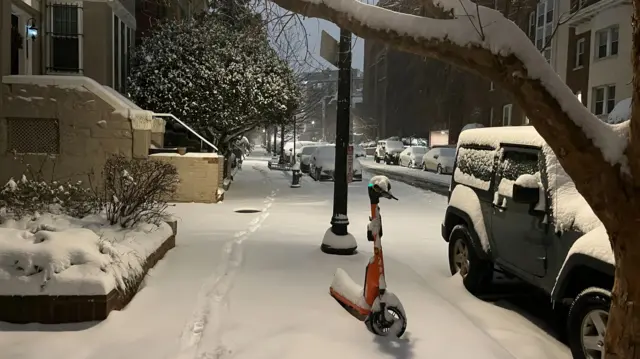 Street covered in snow with cars and a scooter left with a thick covering of snow