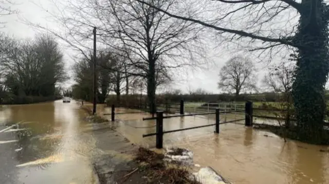 Brown flood water is going across a small bridge and onto a road. There is a car in the distance driving through it.