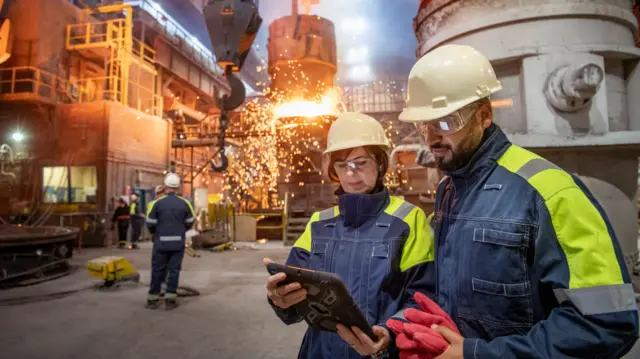 A man and a woman in white helmets and factory gear consult a tablet in the woman's hands while two other workers observe steel pour at the back. They all are on the main factory floor of a steelwork factory