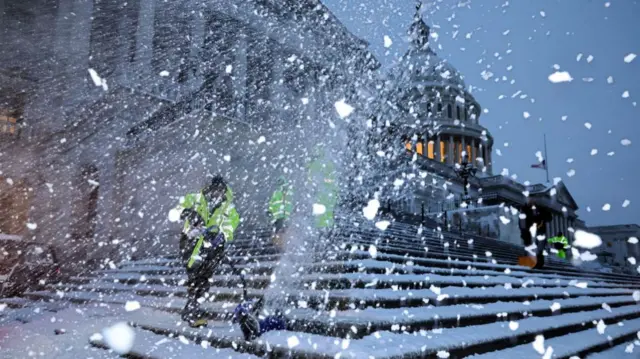 Two crews clear snow from the steps in front of the US Capitol, with snow and ice visible in the foreground