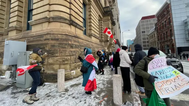 People wearing and waving Canadian flags demonstrate outside the PM's office. One holds a sign saying 'Trudeau's legacy: lawless Canada and show trials'