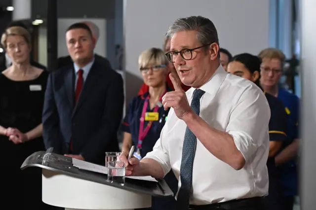 Keir Starmer, in a white shirt and navy tie, points his finger to the audience as Wes Streeting looks on from behind