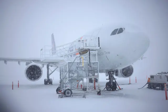 A plane sits on a runway covered in snow, as snow falls around it, with scaffolding at the side of the plane