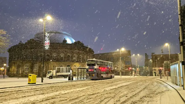 A snowy scene of a bus on the road outside Leeds Corn Exchange