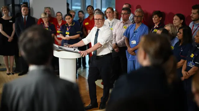 Starmer stands at a white podium with dozens of healthcare workers behind him