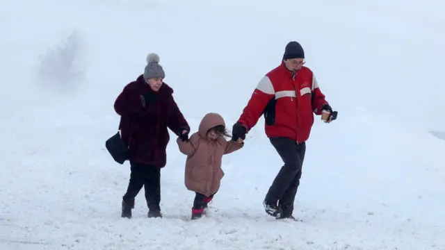 A woman wearing a coat and hat, holds hands with a girl in a pink jacket, who is holding a man's hand who is wearing a red jacket and black hat. They are walking through snow.