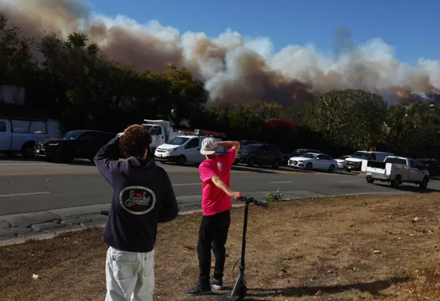 People watch as others drive out of harm’s way as the Palisades Fire burns amid a powerful windstorm