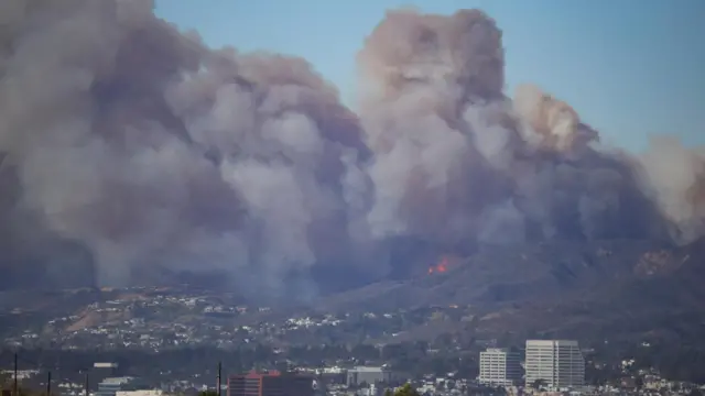 Smoke rises from a wildfire burning on hills near Pacific Palisades, with homes and buildings in the foreground and very close to the fire.