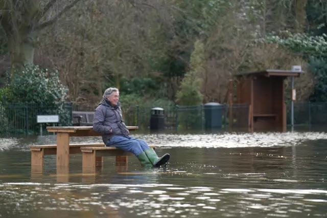 A man sits at a picnic bench and lifts his feet just above the flood water surrounding him