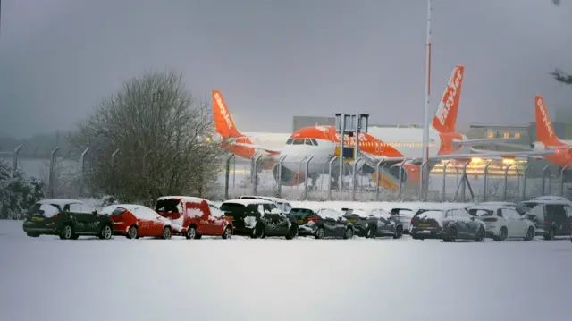 Two easyjet planes on a snowy runway with snow-covered cars in the car park in front