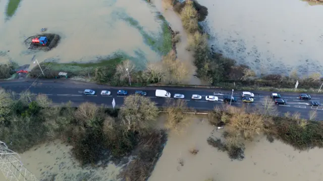 Flooded fields in Glen Parva, Leicestershire. A drone shot shows a queue of cars lined up along the road which is surrounded by flood water