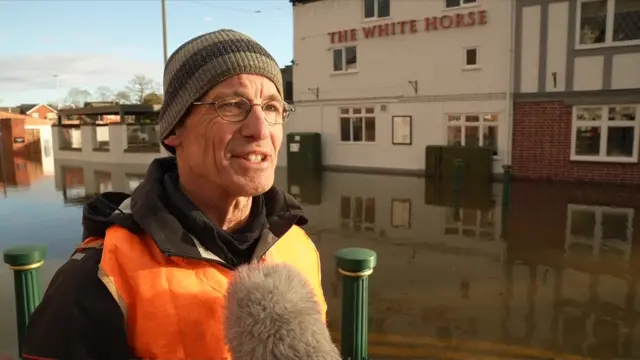 Simon Austin wearing an orange high-vis vest in front of a flooded pub, The White Horse