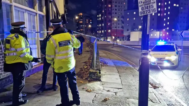 Police tape blocks off access to a main road in London and a person on the other side of the tape is talking with officers in reflective vests.