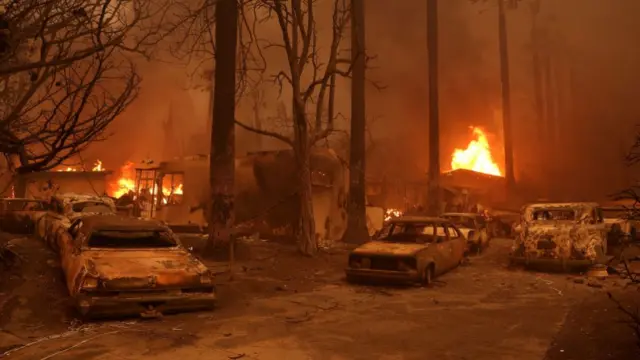 Burned cars sit parked in front of a burning home as the Eaton Fire moves through the area on January 08, 2025 in Altadena, California.