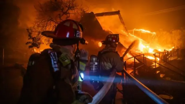 Two firefighters in uniform holding a hosepipe spray water toward huge flames from the Palisades Fire.