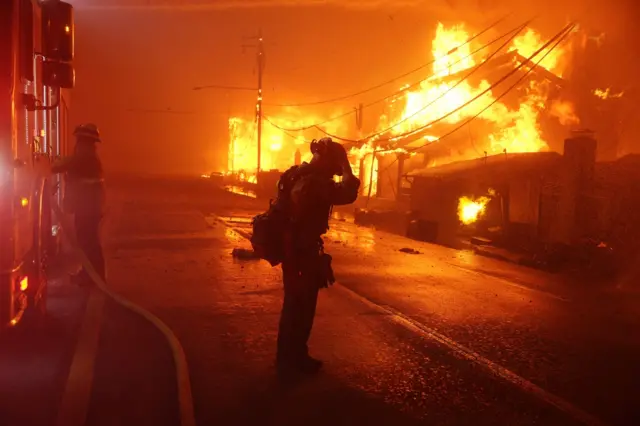 A firefighter touches his head in front of blazing buildings